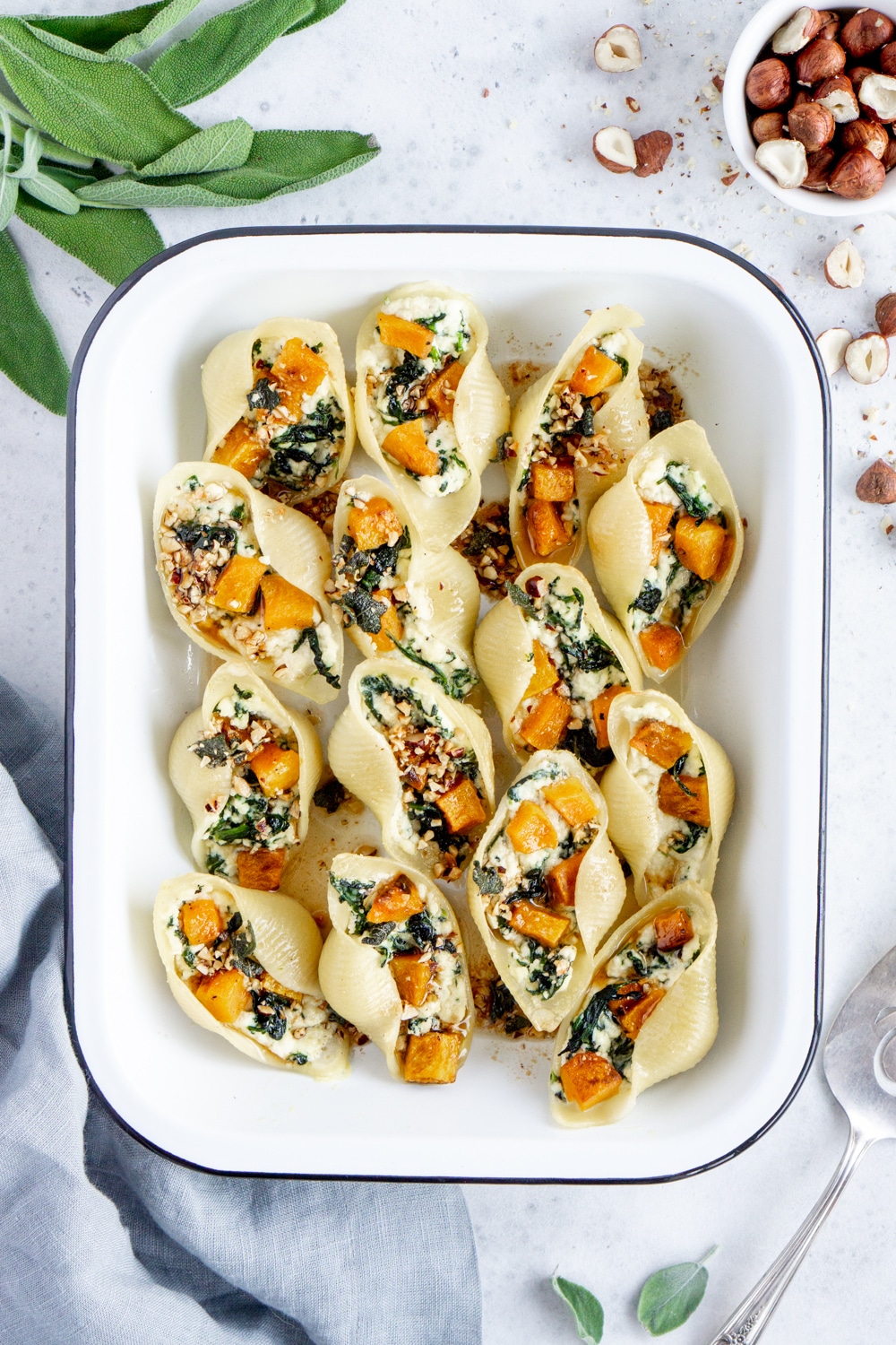 Cooked stuffed pasta shells in a baking dish surrounded by fresh sage and a gray napkin