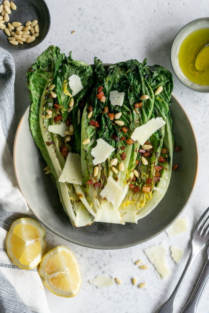Grilled romaine salad in a bowl surrounded by lemons. pine nuts and vinaigrette