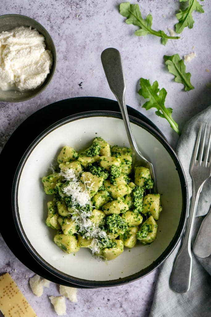 Bowl of cooked ricotta gnocchi with arugula pesto and arugula and parmigiano sprinkled around the scene