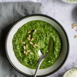 White bowl of arugula pesto topped with pine nuts, and a spoon in the bowl