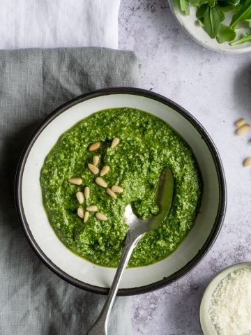White bowl of arugula pesto topped with pine nuts, and a spoon in the bowl