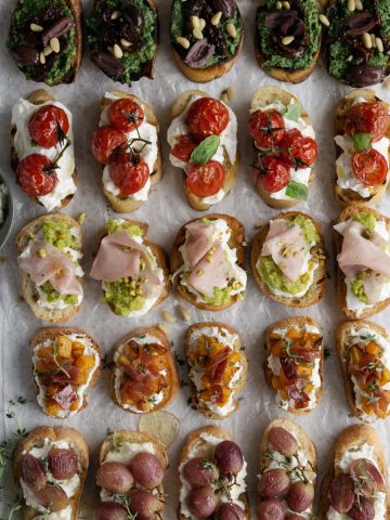 Five different types of crostini arranged on parchment paper with a knife and bowl of ricotta on either side