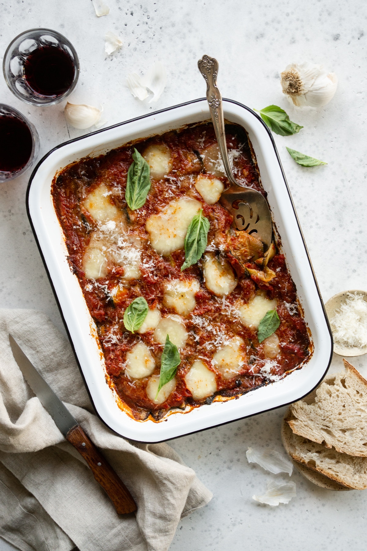baking dish of zucchini parmigiana surrounded by wine glasses, fresh basil and bread