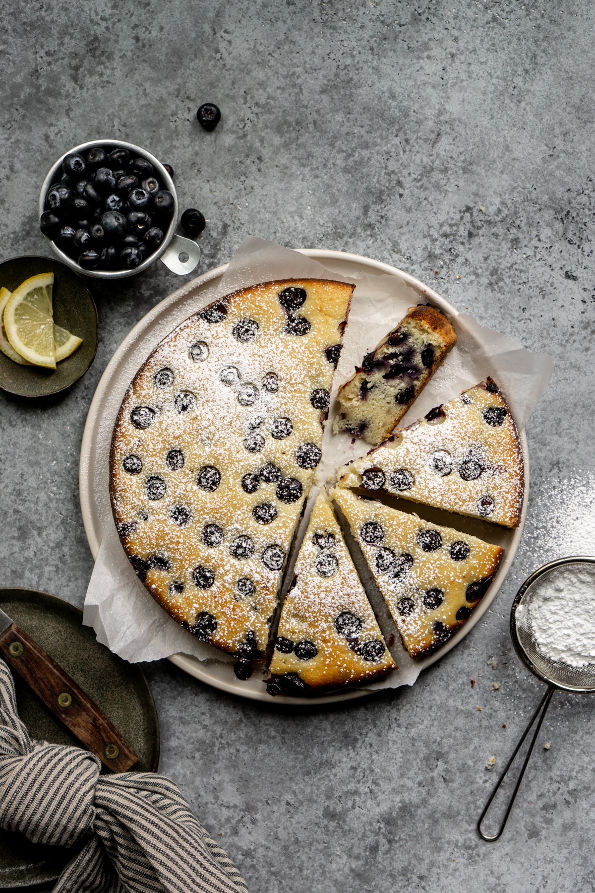 Blueberry ricotta cake in a dish surrounded by a cup of blueberries and lemon wedges
