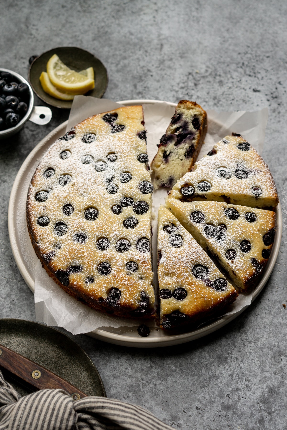 Blueberry ricotta cake in a plate topped with powdered sugar and a dish of lemon slices in the background
