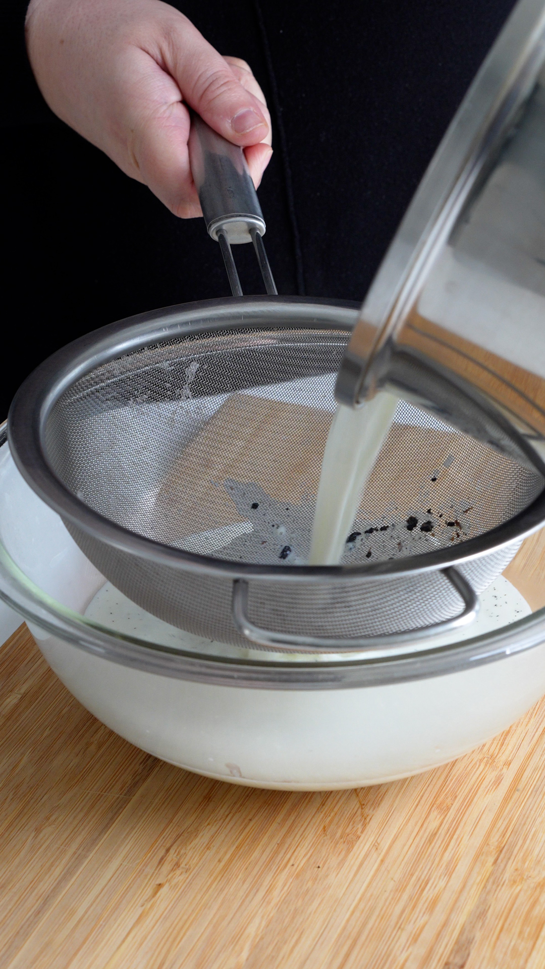 Milk mixture with vanilla beans and seeds being strained into a glass bowl with a colander