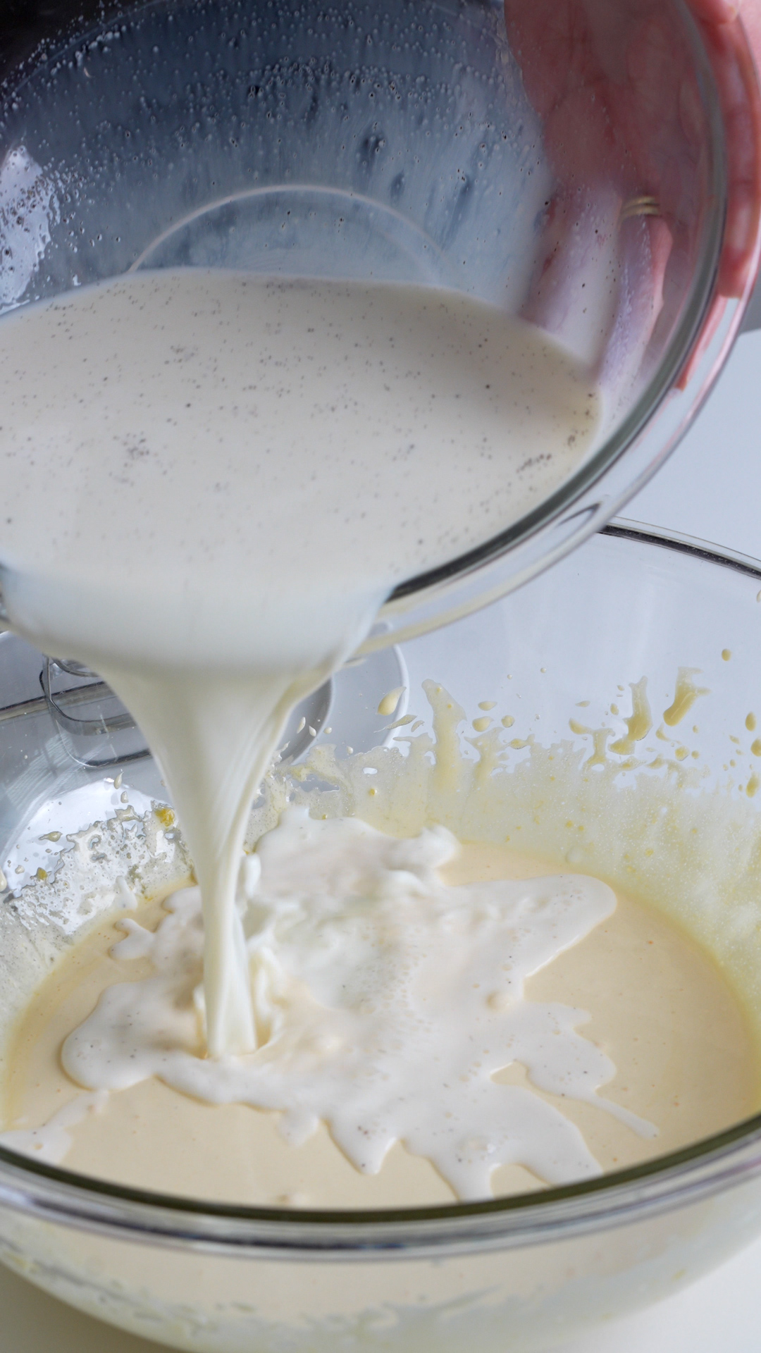 Strained milk mixture being poured into a bowl with whisked egg yolks and sugar