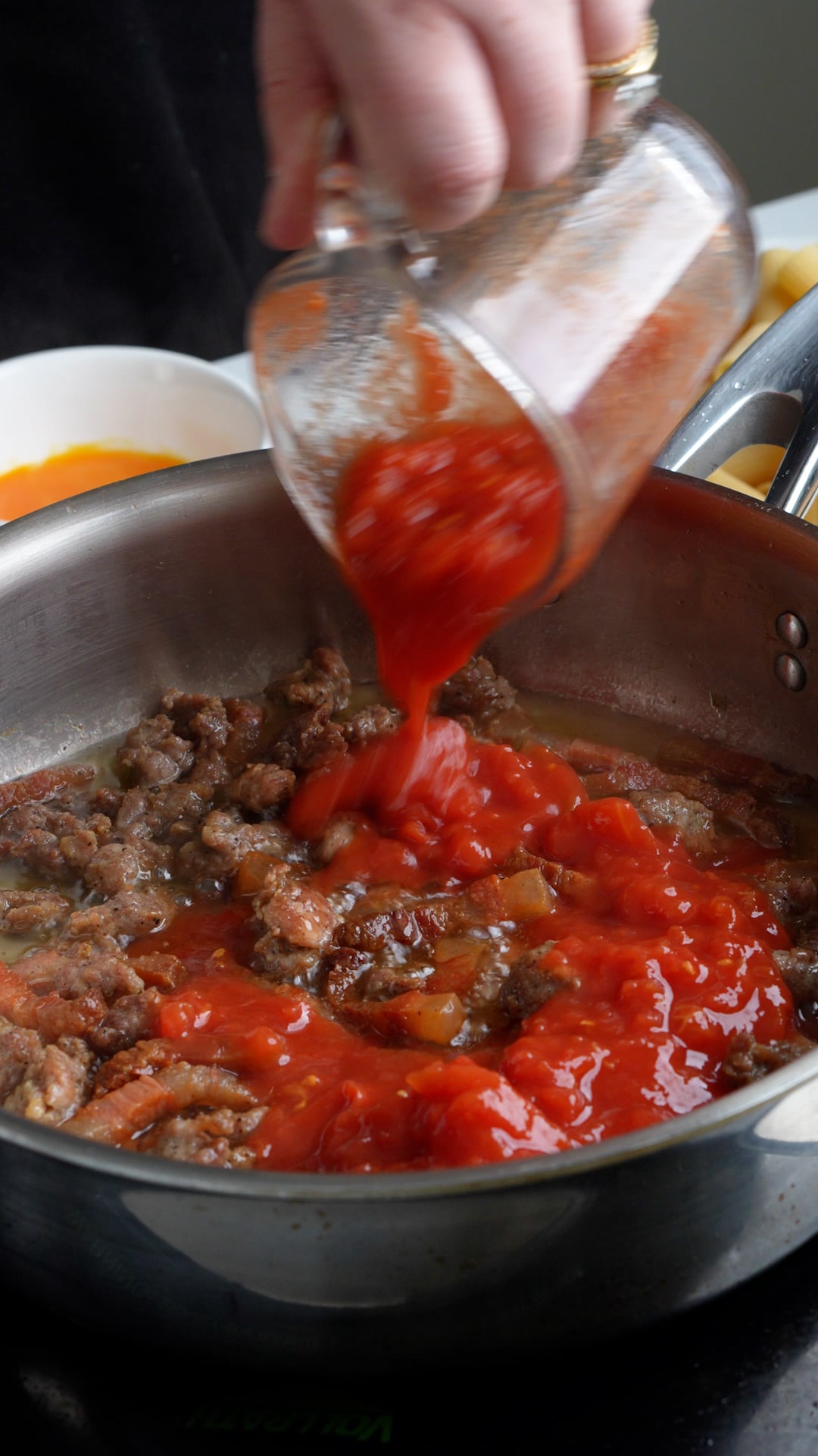 Canned tomatoes being poured into a pan with sausage and guanciale in it