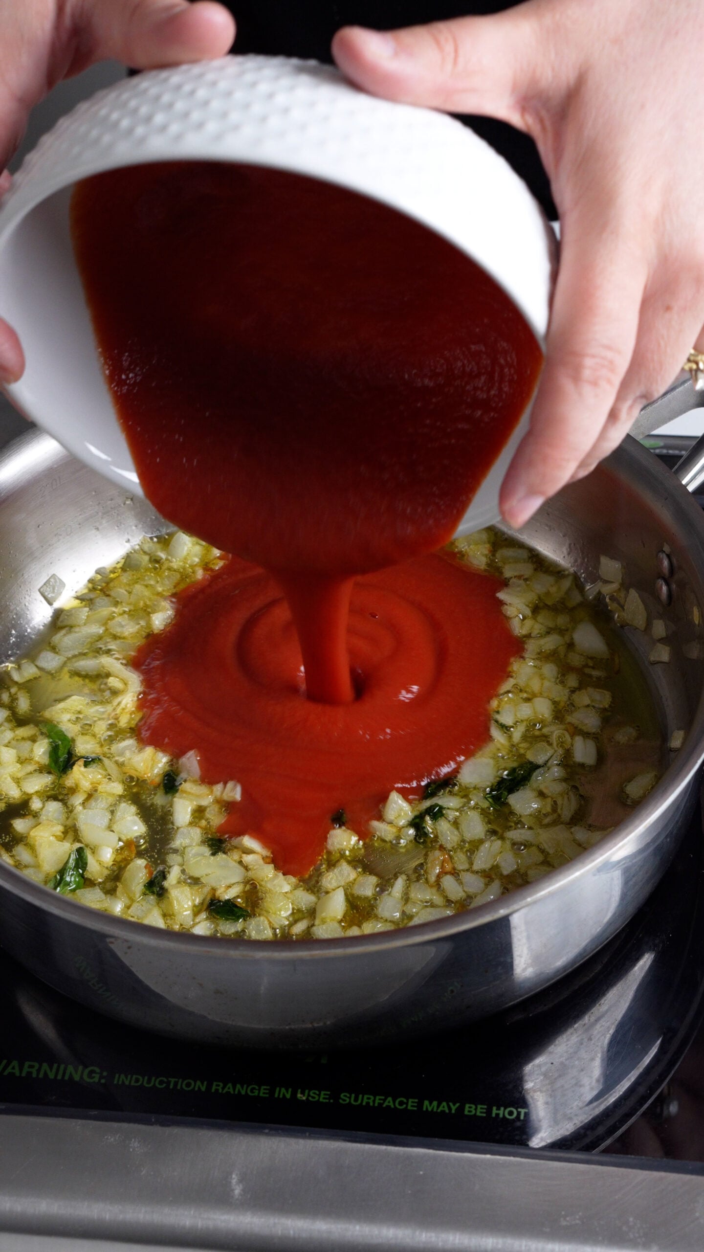 Tomato passata being poured into a pan with olive oil, cooked onion and basil
