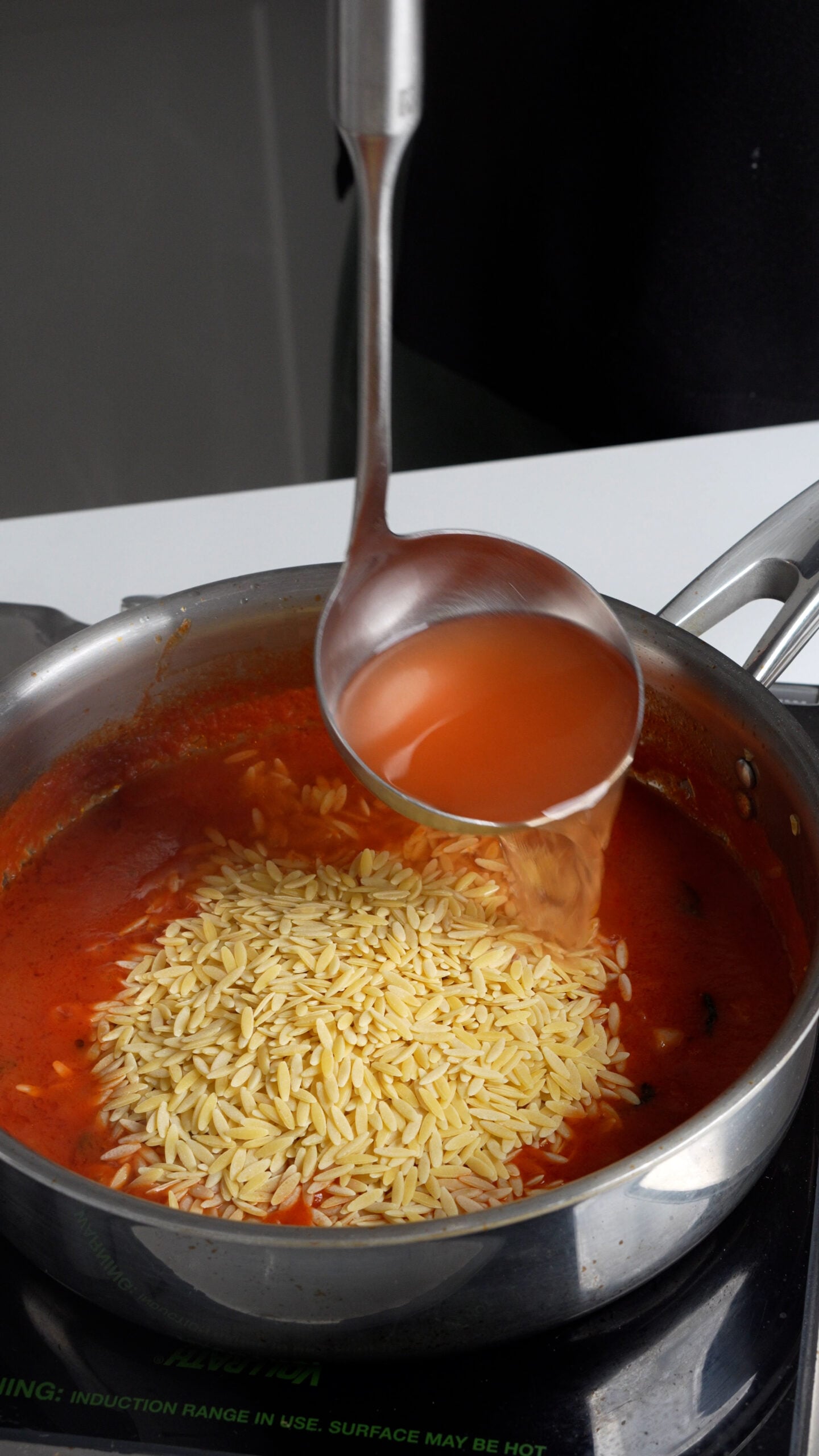 Tomato broth being ladled into a pan containing tomato sauce and pastina