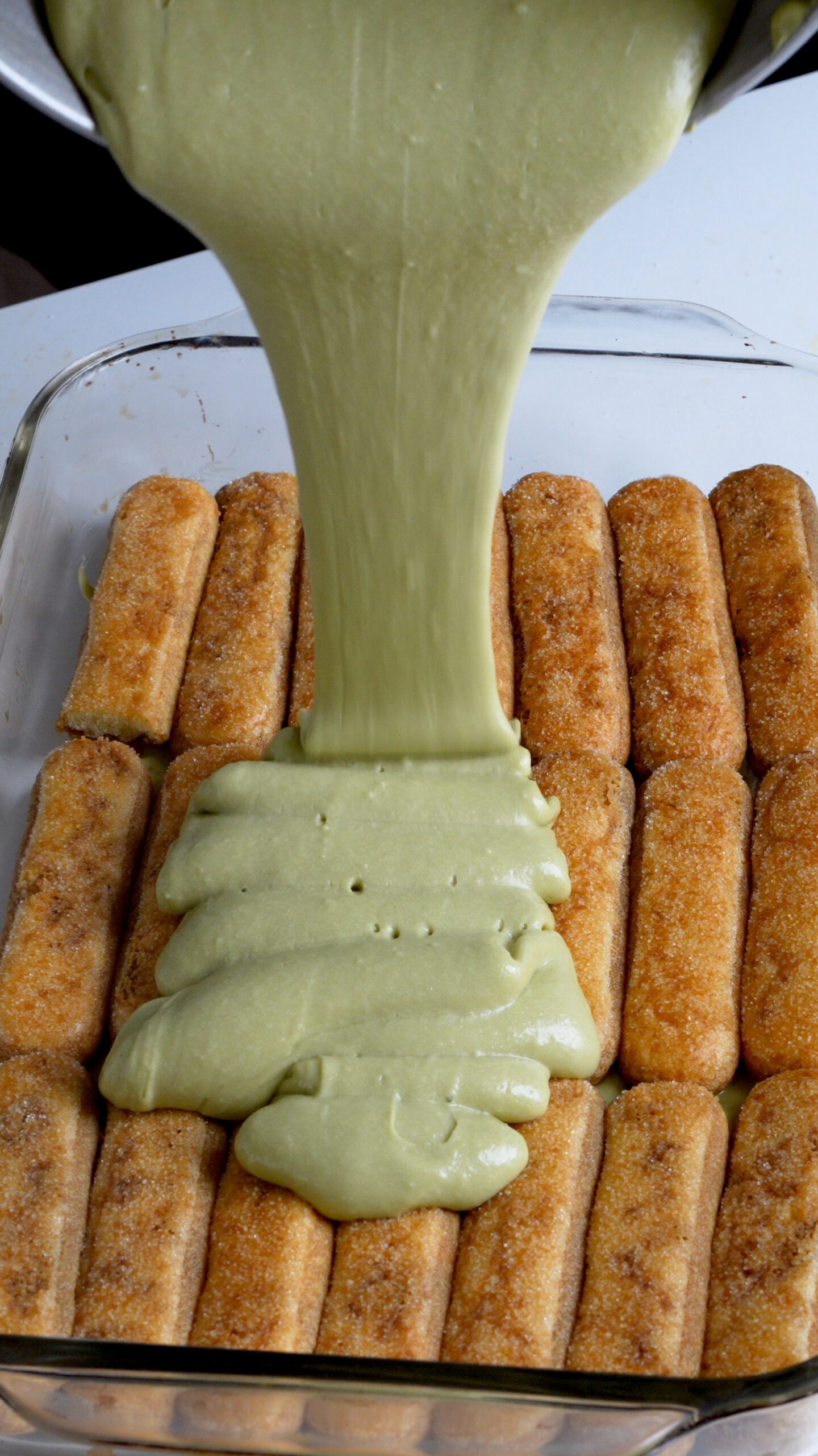 Pistachio tiramisu cream being poured onto a layer of espresso-soaked ladyfinger cookies