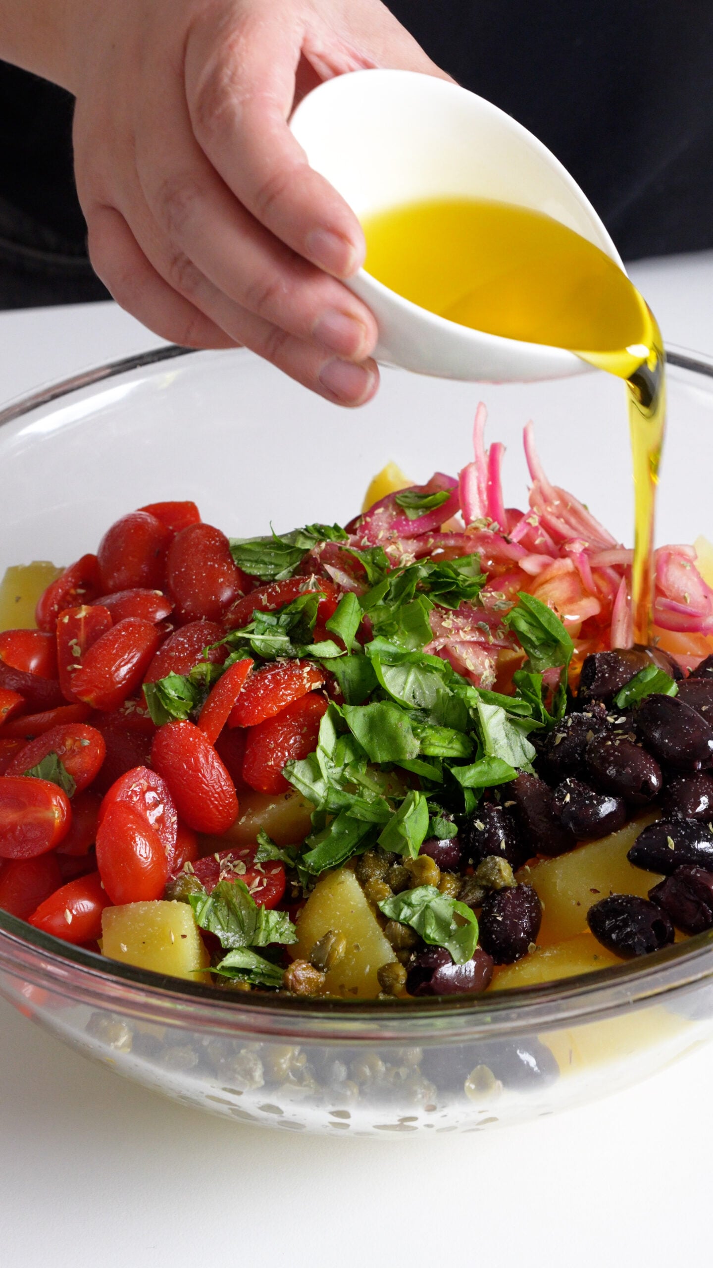 Olive oil being poured onto bowl of cubed potatoes, tomatoes, pickled onions, olives, capers and basil