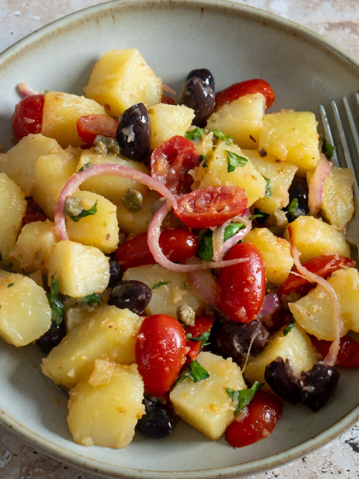 Italian potato salad in a bowl with onions, tomatoes, olives, capers, basil and oregano