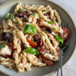 Italian pasta salad in a plate topped with sun dried tomatoes, grape tomatoes, olives, basil, mozzarella and tuna