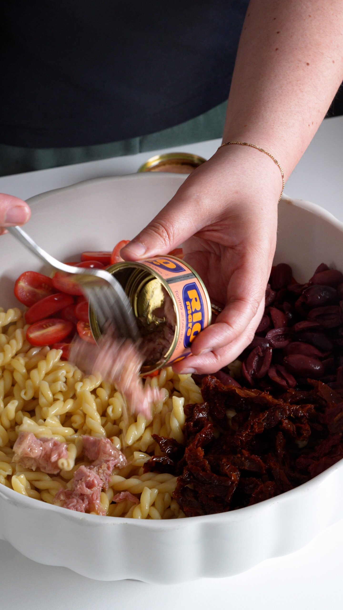 Canned tuna being placed into a bowl with cooked pasta, fresh tomatoes, sun dried tomatoes and olives
