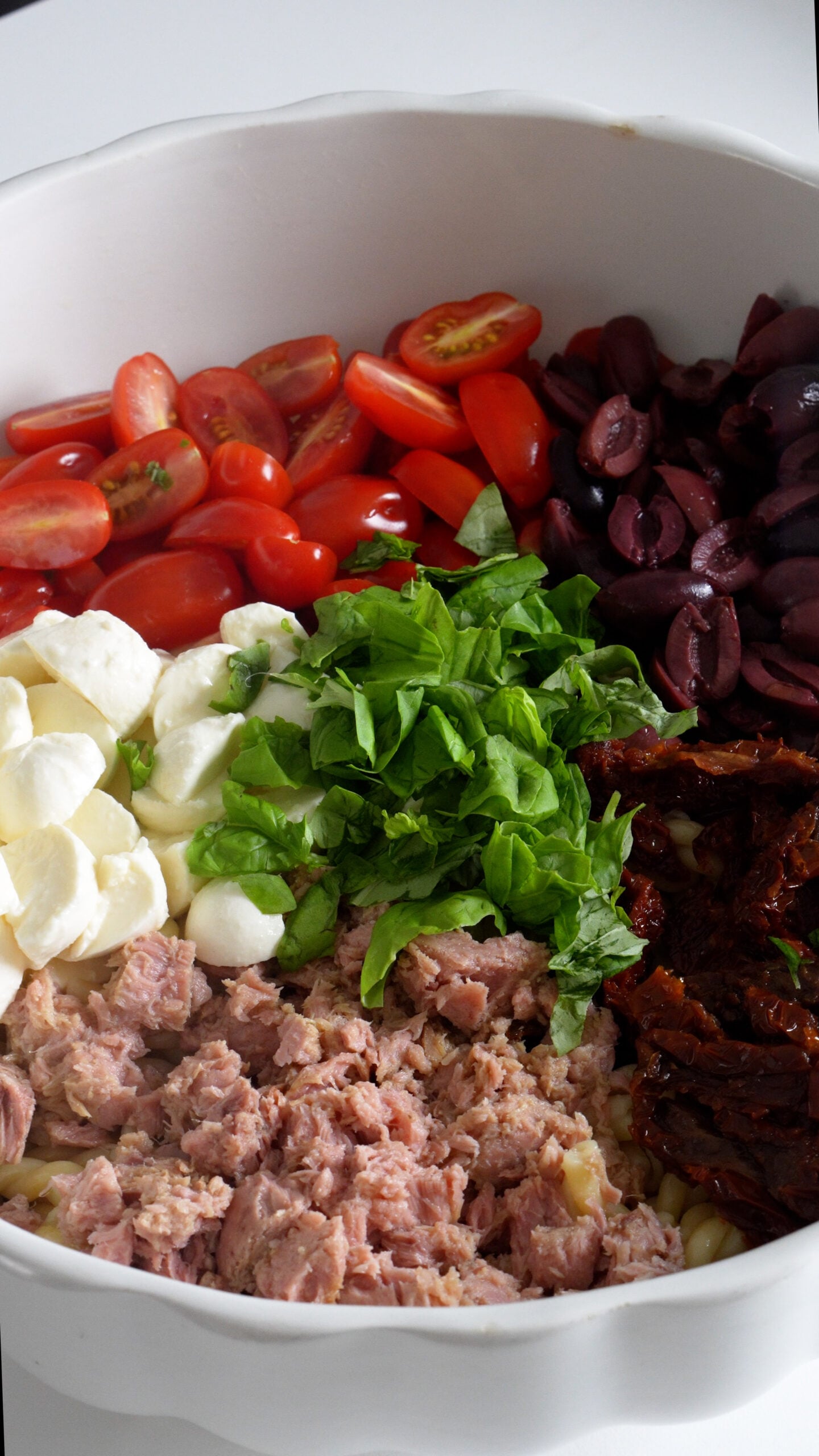Pasta salad ingredients in a bowl before being mixed - olives, sun dried tomatoes, canned tuna, bocconcini, fresh tomatoes and basil